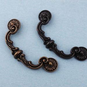 May include: Two ornate cabinet pulls. One is bronze-toned, the other is black. Each pull features a floral design on the circular end pieces and a curved handle with decorative flourishes. The pulls are set against a light blue background.