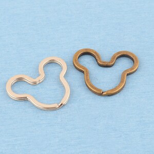 May include: Two metal split rings shaped like mouse heads. One ring has a shiny silver-tone finish, and the other ring has a matte bronze-tone finish. Both rings feature a circular main section with two smaller circular ears at the top.