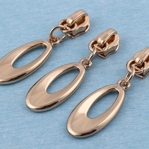 May include: Three gold-toned zipper pulls with oval-shaped charms. Each pull has a metal loop at the top.