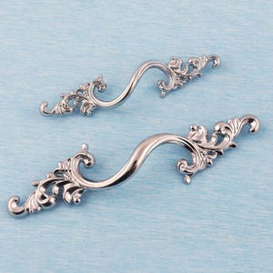 May include: Two silver-colored, ornate cabinet handles with a curved design. Each handle features decorative scrollwork at both ends, adding a touch of elegance. The handles are set against a light blue background, highlighting their polished finish.