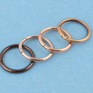 Puede incluir: Cuatro anillas metálicas circulares con resorte dispuestas en línea diagonal. Las anillas cuentan con un mecanismo de apertura con bisagra y vienen en cuatro acabados metálicos distintos: bronce oscuro, oro rosa pulido, oro claro y tono bronce.