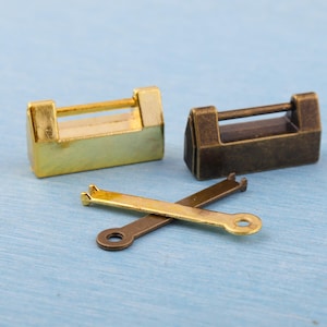 May include: Two small metal padlocks, one gold and one bronze, with matching keys. The gold padlock has a rectangular body and a handle. The bronze padlock has a similar design. The keys are crossed, one gold and one bronze.