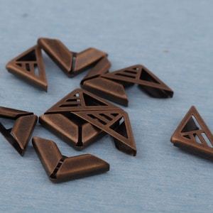 May include: A collection of bronze-colored metal corner protectors. Each piece is triangular with a decorative geometric cutout design. The protectors are scattered on a light blue surface, showcasing their aged, vintage appearance.