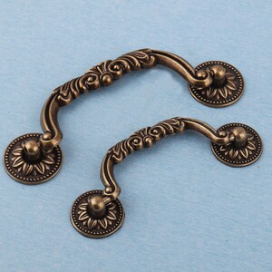 Może przedstawiać: Dwa antyczne uchwyty do szuflad w kolorze brązu. Każdy uchwyt ma zakrzywiony uchwyt z ozdobnymi detalami kwiatowymi i jest przymocowany do okrągłej podstawy z motywem słonecznika. Uchwyty są umieszczone na jasnoniebieskim tle.