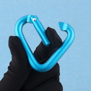May include: A bright blue heart-shaped carabiner with a smooth metallic finish. The carabiner features a spring-loaded gate mechanism that opens inward, allowing for the attachment of items. The frame is constructed from a single piece of metal.