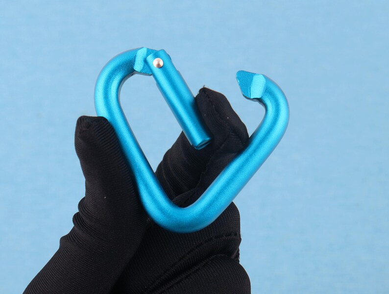 May include: A bright blue heart-shaped carabiner with a smooth metallic finish. The carabiner features a spring-loaded gate mechanism that opens inward, allowing for the attachment of items. The frame is constructed from a single piece of metal.
