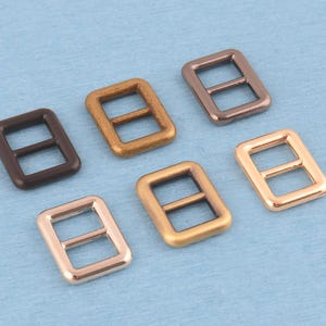 May include: Six rectangular metal strap adjusters or sliders arranged in two rows. The set includes various metallic finishes, specifically matte dark brown, antique brass, polished gunmetal, rose gold, brushed gold, and bright gold tones.