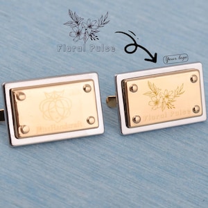 May include: Two rectangular cufflinks with a gold-coloured centre and silver-coloured border. Each cufflink has an engraved floral design and the text "Floral Pulse" and "MissHandcraft". The cufflinks are on a blue surface.