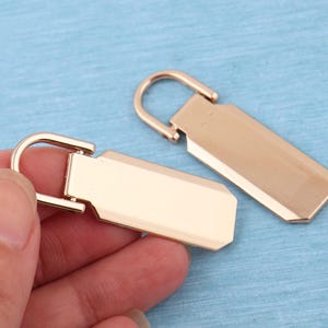 Peut inclure: Deux tirettes de fermeture &eacute;clair en m&eacute;tal dor&eacute; rose de forme rectangulaire avec des coins arrondis. Chaque tirette comporte une boucle semi-circulaire articul&eacute;e sur le dessus pour la fixation. La surface du m&eacute;tal est lisse et brillante.
