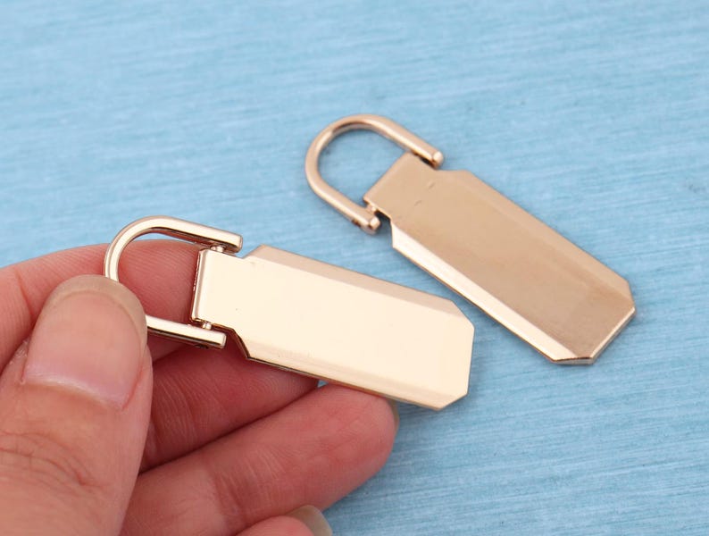 Peut inclure: Deux tirettes de fermeture &eacute;clair en m&eacute;tal dor&eacute; rose de forme rectangulaire avec des coins arrondis. Chaque tirette comporte une boucle semi-circulaire articul&eacute;e sur le dessus pour la fixation. La surface du m&eacute;tal est lisse et brillante.