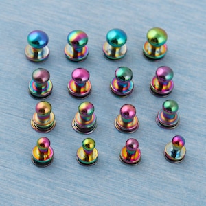 May include: Sixteen iridescent metal buttons with a domed top and a flat base are arranged on a light blue surface. The buttons display a range of colors, including blues, greens, pinks, and yellows, creating a rainbow effect.