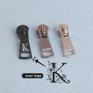 May include: Three metal zipper pulls in black, silver, and bronze. The black pull features a floral "K" design. The silver pull is stamped "Hand Made". The image includes a logo design with the text "your logo".