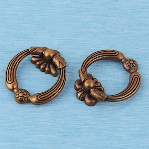 May include: Two circular bronze-toned metal drawer pulls featuring a decorative floral-inspired mounting plate at the top and a small embossed flower detail at the bottom, with grooved lines running along the sides of the ring-shaped handle.