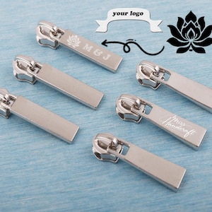May include: Silver metal zipper pulls with engraved text and a lotus flower design. The pulls are rectangular with a rounded top and are scattered on a blue surface. Some pulls have the text "M&J" and "Miss Handcraft".
