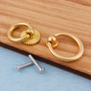 Op de afbeelding: Twee goudkleurige kastgrepen met ringgrepen en decoratieve achterplaten. De grepen worden getoond met twee zilveren schroeven, klaar voor installatie. De hardware is geplaatst op een houten oppervlak.