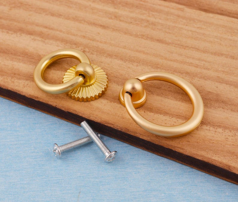 Op de afbeelding: Twee goudkleurige kastgrepen met ringgrepen en decoratieve achterplaten. De grepen worden getoond met twee zilveren schroeven, klaar voor installatie. De hardware is geplaatst op een houten oppervlak.