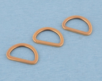 May include: Three metallic D-rings with a smooth, copper-toned finish. Each piece features a curved top edge and a straight bottom bar, forming a classic semi-circular shape. These hardware components are arranged in a diagonal pattern.