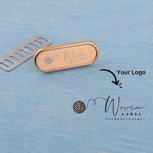Può includere: Un'etichetta metallica color oro con la scritta "Woven Label" e una grafica a forma di gomitolo. Un pezzo di metallo separato è accanto all'etichetta. L'etichetta è su una superficie azzurra.