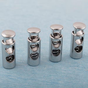 May include: Four shiny, silver cord locks with a cylindrical body and a rounded top. Each lock has a spring mechanism visible through the side openings. The background is a light blue surface.