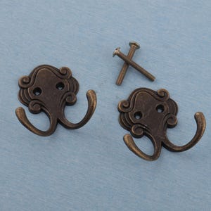 Könnte beinhalten: Zwei antike, bronzefarbene Doppel-Kleiderhaken mit dekorativen Schnörkeln und zwei Schrauben. Die Haken befinden sich auf einer hellblauen Oberfläche. Die Haken sind zum Aufhängen von Mänteln, Hüten oder anderen Gegenständen bestimmt.