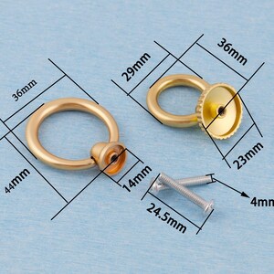 Op de afbeelding: Twee goudkleurige ringtrekkers met bevestigingsmateriaal. De ringtrekkers hebben een diameter van 3,6 cm en zijn 4,4 cm hoog. De ring zelf heeft een diameter van 2,9 cm. De basis heeft een diameter van 2,3 cm en is 1,4 cm hoog. Schroeven zijn 2,45 cm lang en 4 mm in diameter.