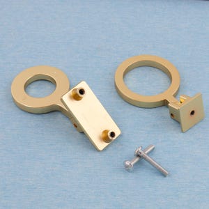 Puede incluir: Dos tiradores dorados para armarios con dise&ntilde;o circular y placas de montaje rectangulares. Tambi&eacute;n se incluyen dos tornillos plateados. La ferreter&iacute;a se encuentra sobre un fondo azul claro.