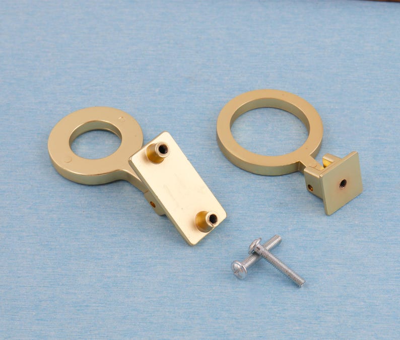 Puede incluir: Dos tiradores dorados para armarios con dise&ntilde;o circular y placas de montaje rectangulares. Tambi&eacute;n se incluyen dos tornillos plateados. La ferreter&iacute;a se encuentra sobre un fondo azul claro.