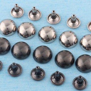 May include: Assortment of silver and black metal dome-shaped studs with a pointed post. The studs are arranged in a random pattern on a blue surface.