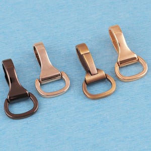 May include: Four metal swivel snap hooks with D-rings, arranged in a row. The hardware features four distinct metallic finishes: a dark gunmetal, a light silver, a brushed bronze, and a polished rose gold, each with a smooth, curved design.