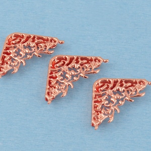 May include: Three rose gold-coloured metal corner protectors featuring an intricate, openwork filigree pattern. Each piece is triangular in shape with curved edges and a polished, reflective surface finish, arranged in a diagonal row.