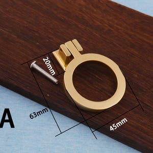 Puede incluir: Un tirador de armario dorado con un asa circular, que mide 4,5 cm de di&aacute;metro, 6,3 cm de largo y 2 cm de alto. Incluye un tornillo plateado para la instalaci&oacute;n. La letra "A" es visible. El tirador se muestra sobre una superficie de madera oscura.