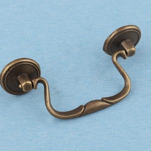 May include: A brass-toned metal drawer pull featuring a curved, swinging handle attached to two circular mounting plates. The hardware has a weathered, darkened finish with visible highlights on the edges and a smooth, contoured shape.