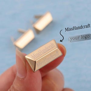 Puede incluir: Primer plano de herrajes metálicos rectangulares dorados con un poste en la parte posterior. El texto "MissHandcraft" está grabado en la parte delantera. Los herrajes se utilizan probablemente para bolsos, carteras u otros proyectos de artesanía.