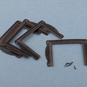 May include: A set of antique bronze-coloured metal corner brackets, each with a decorative curved edge and pre-drilled holes for screws. Several brackets are stacked, with a few screws scattered nearby. The brackets are designed for furniture or craft projects.