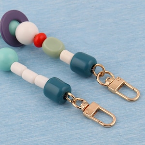 May include: A colorful beaded bag strap featuring a variety of beads in shades of purple, teal, white, red, and green. The strap has gold-colored clasps on each end for easy attachment to a bag.