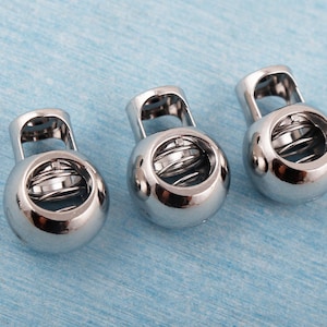 Puede incluir: Tres cierres de cordón plateados y brillantes, cada uno con un mecanismo de resorte visible dentro del cuerpo esférico. Tienen un pequeño lazo rectangular en la parte superior. Fondo azul claro texturizado.