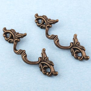 Puede incluir: Dos tiradores de cajón antiguos de color bronce con diseños florales ornamentados y en espiral. Los tiradores tienen forma de asa curva y están sobre un fondo azul claro. Estas piezas de ferretería decorativas añaden un toque vintage.