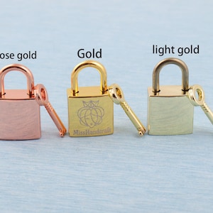 May include: Three miniature padlocks in rose gold, gold, and light gold, each with a key. The gold padlock has an engraved design and the text "MissHandcraft". The background is a light blue surface.