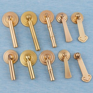 以下が含まれることがあります： さまざまなデザインの真鍮製キャビネットプル9つのコレクション。プルは丸いバックプレートと装飾的なハンドルを備えており、竹風のディテールや装飾的な湾曲した端を持つものもあります。ハードウェアは水色の表面に配置されています。