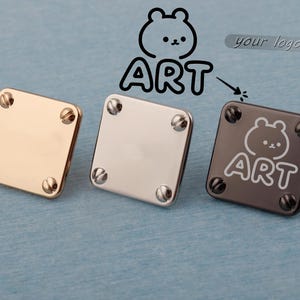 Könnte beinhalten: Drei quadratische Metallbeschläge in Gold, Silber und Dunkelgrau. Jedes hat vier Schrauben an den Ecken. Das dunkelgraue Stück zeigt eine Bärengrafik und das Wort "ART". Der Hintergrund ist hellblau.