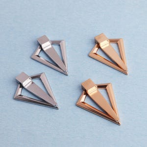 May include: Four metal clasps, two silver and two gold. Each clasp features a rectangular top and a triangular frame. The clasps are displayed on a light blue surface.