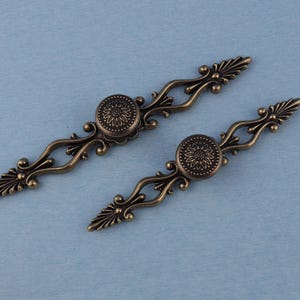 May include: Two antique bronze cabinet pulls. Each pull features a central circular medallion with a floral design, flanked by ornate, curved arms and decorative end pieces. The pulls are set against a light blue background.