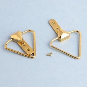 May include: Two gold cabinet pulls with a geometric design. Each pull has a triangular handle and a faceted centre piece. Two small screws are included. The pulls are shown on a light blue surface.
