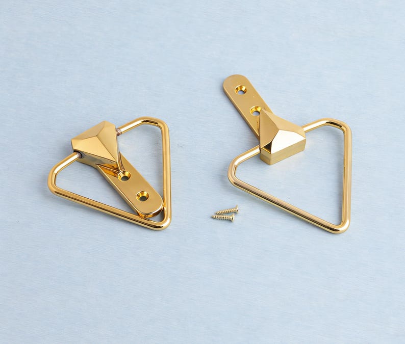 May include: Two gold cabinet pulls with a geometric design. Each pull has a triangular handle and a faceted centre piece. Two small screws are included. The pulls are shown on a light blue surface.