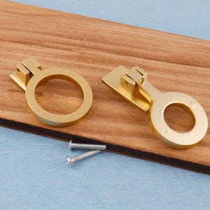 Puede incluir: Dos tiradores dorados para armarios con dise&ntilde;o de anillo, sobre una superficie de madera. Los tiradores van acompa&ntilde;ados de dos tornillos plateados. El herraje est&aacute; dise&ntilde;ado para proyectos de mobiliario y decoraci&oacute;n del hogar.