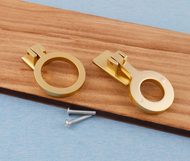 Puede incluir: Dos tiradores dorados para armarios con dise&ntilde;o de anillo, sobre una superficie de madera. Los tiradores van acompa&ntilde;ados de dos tornillos plateados. El herraje est&aacute; dise&ntilde;ado para proyectos de mobiliario y decoraci&oacute;n del hogar.