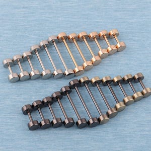 Puede incluir: Una variedad de pendientes de botón con diferentes acabados metálicos, incluyendo plata, oro, oro rosa y metal de cañón. Los pendientes están dispuestos en filas sobre una superficie azul claro, mostrando su diseño en forma de pesa.