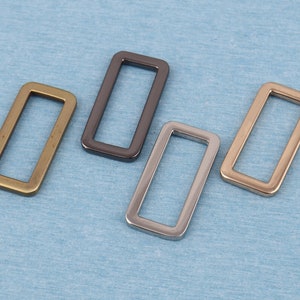 May include: Four metal rectangular rings in different finishes: gold, silver, black nickel, and brass. The rings are all the same size and shape.