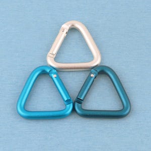 May include: Three triangular carabiners arranged in a pyramid formation. One carabiner is a matte silver color, while the other two are shades of blue, with one being a bright cyan and the other a deeper teal, each featuring a spring-loaded gate.