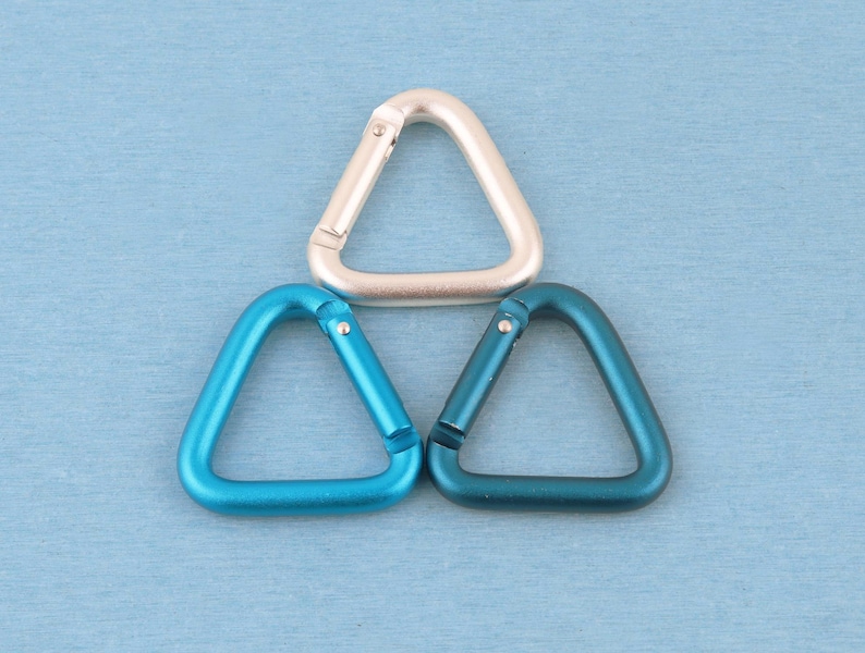 May include: Three triangular carabiners arranged in a pyramid formation. One carabiner is a matte silver color, while the other two are shades of blue, with one being a bright cyan and the other a deeper teal, each featuring a spring-loaded gate.